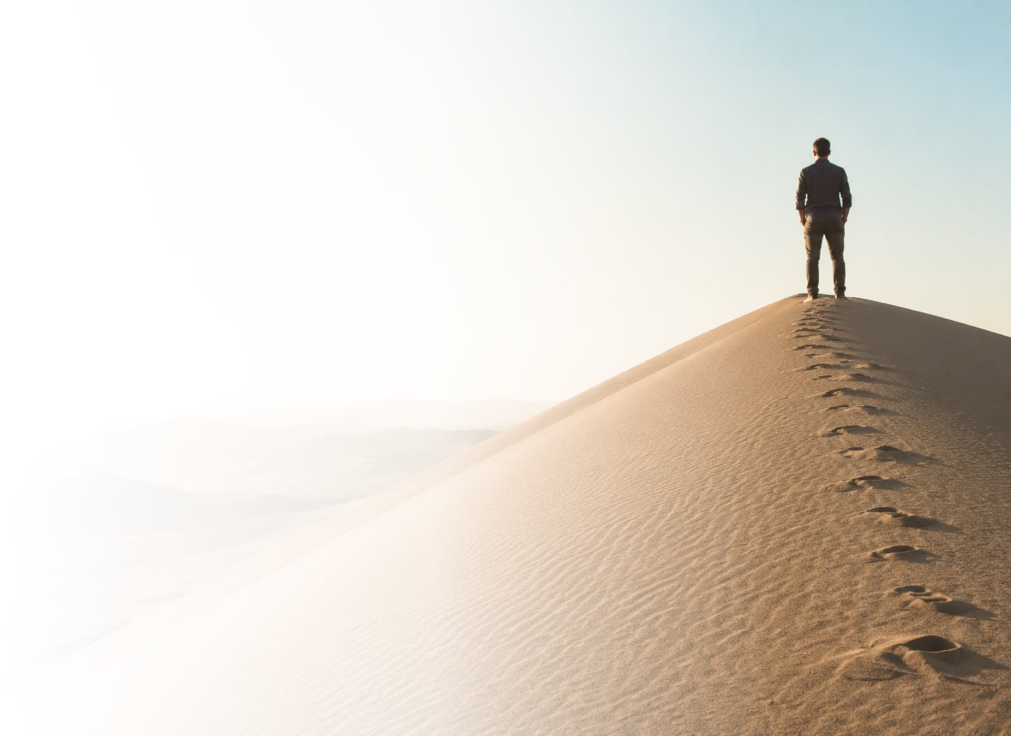 A man walking alone along the crest of a sand dune, his footprints trailing behind him