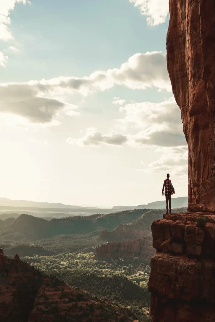 A hiker standing at the edge of a red-rock cliff, looking out over a wide valley